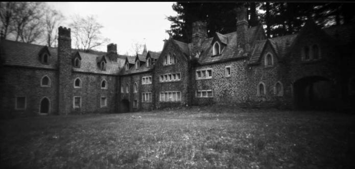 The image depicts a black and white photograph of a large, old stone building resembling a castle or manor. The structure features several gabled roofs and multiple chimneys, giving it a historic and gothic appearance. The facade includes numerous arched windows, each framed in stone, with some windows grouped in clusters. The building is L-shaped, creating a courtyard effect with the grassy foreground. The right side of the structure includes a large arched entryway leading to the courtyard. Bare trees and tall evergreens are visible behind the building, adding to the moody atmosphere.