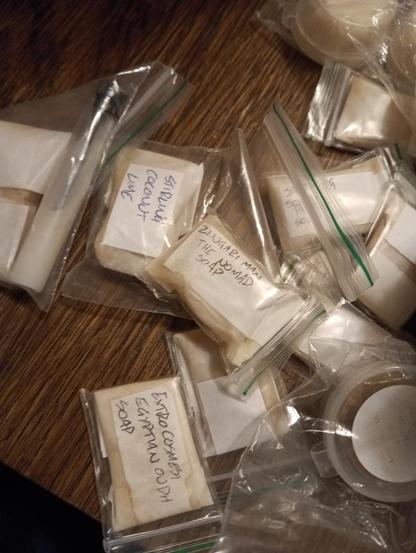The image shows several small, individually wrapped soap samples in clear plastic bags with handwritten labels. The labels indicate different scents such as "Zingari Man The Nomad Soap," "Extra Cosiness Egyptian Oudh Soap," and "Stirling Coconut Lime." Some soaps are stacked or overlapping, and a small container with a white lid is also visible. The scene is set on a wooden surface, and the soaps appear to be artisan or speciality shaving soaps.