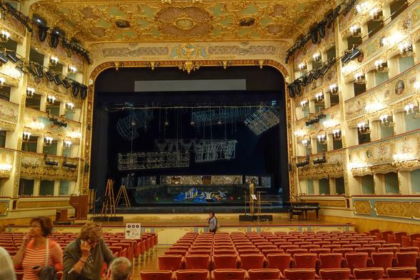 Interior of La Fenice opera house in Venice. The decoration is rococo and lavishly ornate, abundantly gilded, and lit by dozens of wall sconces. Both sides of the theatre are composed of four storeys of boxes, and the proscenium arch of the stage is huge and almost square, nearly the full height and width of the interior.