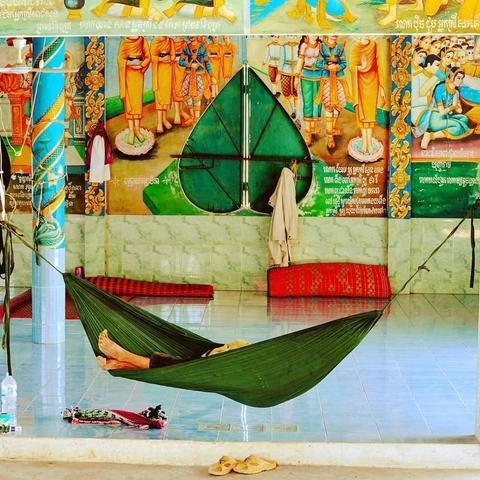 Photo shows a green hammock with a couple of feet sticking out from the person sleeping there. A pair of bright yellow sandals are neatly put under the hammock. It is hanged up in an half open room in a pagoda complex, and the walls are painted with colorful paintings of the Lord Buddhas life and teachings. A big green window cover in the shape of a lotus leaf is visible in the middle. Small red matts are rolled up towards the wall.