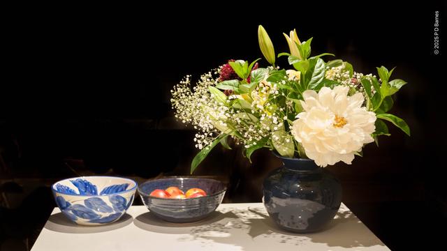 A still life with a vase filled with predominantly white flowers—peonies, baby's breath, lilies—and two bowls, one solid blue filled with apples, and one empty, white with brushed blue leaf pattern.