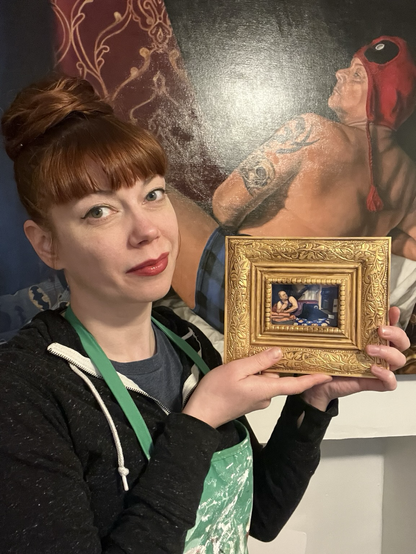 Artist Klaire Lockheart, wearing a splattered green apron, holds up a tiny print of a reproduction of a brodalisque painting in an ostentatious gold frame painting.