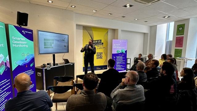Das Bild zeigt Horst Thieme, der während der FUTURE WEEK 2024 im Future Store in Augsburg einen Vortrag hält. Er steht vor einem Publikum und spricht in ein Mikrofon, während er mit einer Hand gestikuliert. Hinter ihm befindet sich eine große Präsentationsfläche mit einer Folie zum Thema Smart City, die über einen Bildschirm angezeigt wird.

Links und rechts von der Bühne stehen große, farbige Infotafeln mit Themen wie „Smartes Gewässer-Monitoring“ und „Smarte Baumbewässerung“, die digitale Innovationen der Stadt Augsburg präsentieren. Im Hintergrund hängt ein großes gelb-schwarzes Banner mit dem Titel der Veranstaltung und einem QR-Code.

Das Publikum besteht aus einer bunt gemischten Gruppe, die aufmerksam zuhört. Einige sitzen in Stuhlreihen, während andere im Stehen folgen. Die Atmosphäre wirkt modern, professionell und innovationsgetrieben – ein idealer Rahmen für den Austausch über digitale Zukunftslösungen für die Stadt.