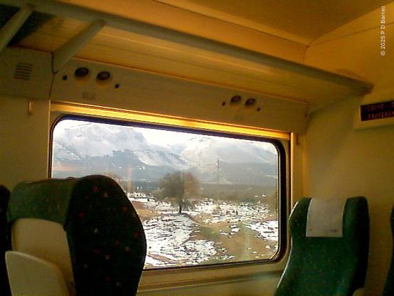 Snow-covered field with olive trees, and a low mountain range behind, seen through the picture window of a high-speed train, with seats and luggage rack in picture. The shot is very low resolution, taken with a 640x480 P910i camera, state of the art at the time.