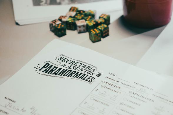 Fotografía a color, plano cerrado de mesa mostrando varios dados de 6 lados en la parte superior, debajo hay una hoja de personaje con el logo del juego de rol "Secretaría de Asuntos Paranormales".