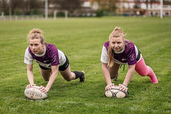 Zwei Rugbyspielerinnen bei einer Aufwärmübung auf dem Platz (Liegestütz auf dem Ball), eine hat ihr gesamtes linkes Bein in einem pinken Gipsverband.