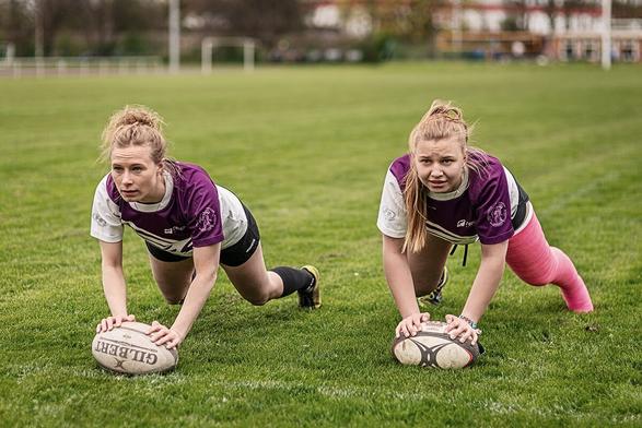 Zwei Rugbyspielerinnen bei einer Aufwärmübung auf dem Platz (Liegestütz auf dem Ball), eine hat ihr gesamtes linkes Bein in einem pinken Gipsverband.