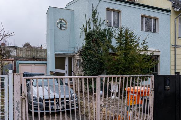 Ein Mercedes-Coupé steht in der Einfahrt eines etwas verwahrlosten Grundstücks. Die ausgeblichene und bewachsene Fassade eines in die Jahre gekommenen Hauses ist zu sehen.