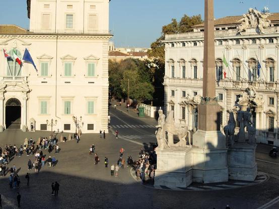 Farbfoto, das Teile des Quirinalspalasts in Rom zeigt, der ab 1583 auf dem Quirinal errichtet wurde und heute als Sitz des italienischen Staatspräsidenten dient. Rechts vorne im Bild der Dioskurenbrunnen mit Obelisk. Auf dem Platz tummeln sich viele Leute.