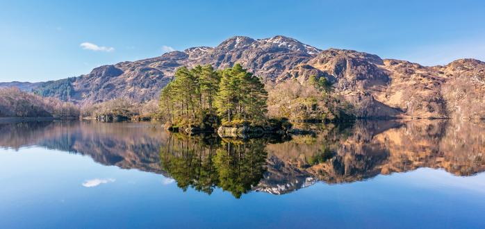 Looking across a mirror-smooth loch, we see an island with evergreen trees sitting in front of distant hills with some snow patches on the tops. The hills and the trees are perfectly reflected in the completely still water, as are the sky and small white clouds, giving a strong sense of symmetry.