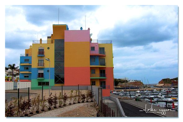 Farbfoto im Querformat. Ein farbenfrohes, mehrstöckiges Gebäude in kubistischer Architektur, das an einem Segelhafen liegt. Die Fassade ist quietschbunt in quadratischen Feldern in den Farben Rosa, Gelb, Orange, Himmelblau und Grün gestrichen. Im Vordergrund befinden sich neu gepflanzte Hecken, die das Gebäude vom Gehweg trennen. Der Himmel ist bewölkt.