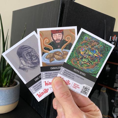 A hand holding three art cards featuring different designs, including a gorilla sketch, a man with a chicken, and a colorful abstract face. A potted plant and electronics are in the background.