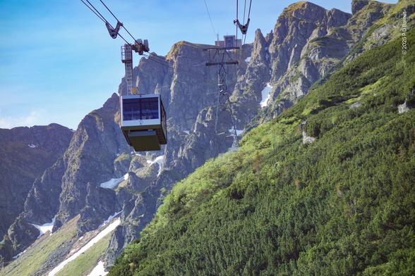 Inside a cable car, hanging over a very steep, partly wooded mountainside, looking up towards the bare rocks at the peak, still holding pockets of snow in shaded clefts. Above are the two sets of cables, and approaching on its downward journey is another large, box-shaped cable car.