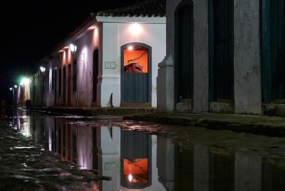 A cobblestone street of a historic center during night time. The middle of the street is filled with water, which reflects the light of the street lamps and the white colonial style buildings with colorful windows and doors.