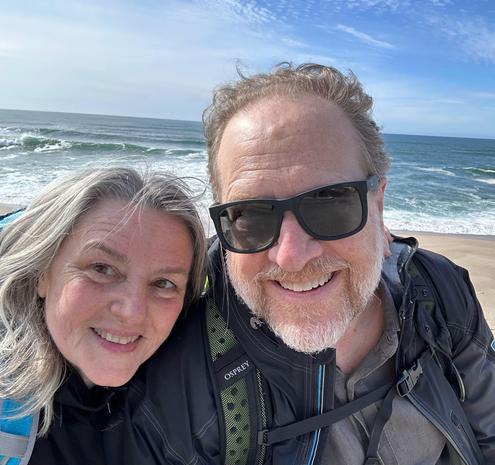 Selfie of a man and a woman in hiking gear smiling at the camera with beach, surf, and blue sky behind them.