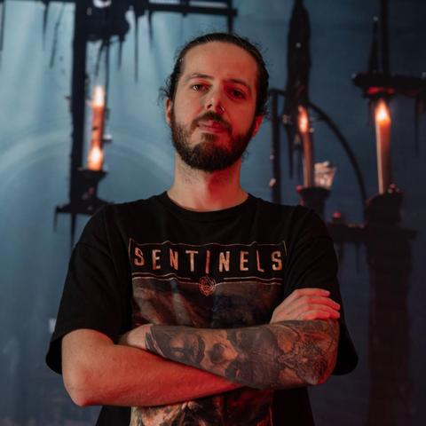 David mit verschränkten Armen und Bandshirt vor einem dunklen Hintergrund mit Kerzen.
