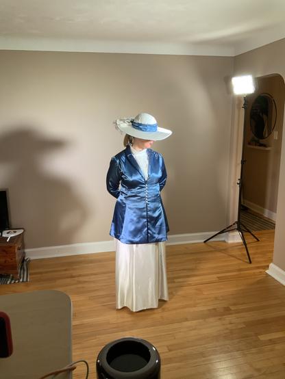 A woman in a blue satin jacket and ivory top and full length skirt with matching wide brimmed white hat stands looking down in a room with dark taupe walls and hardwood floor.