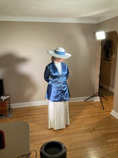 A woman in a blue satin jacket and ivory top and full length skirt with matching wide brimmed white hat stands looking down in a room with dark taupe walls and hardwood floor.