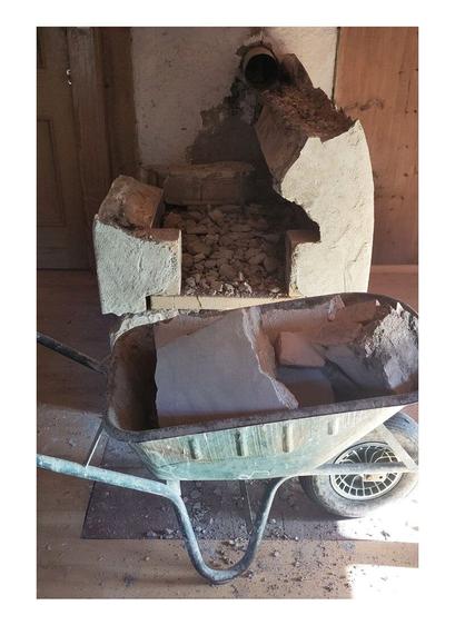 A demolished old oven in pieces of fireclay bricks. Wheelbarrow loaded with brick parts. Wooden old paneling in the background.