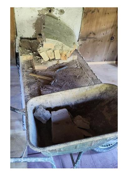 A demolished old oven in pieces of fireclay bricks. Hammer and chisel lie on the remaining oven base. Wheelbarrow loaded with brick parts. Wooden old paneling in the background.
