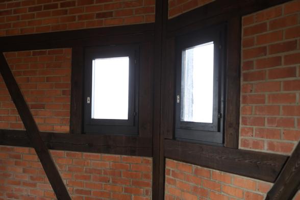 Ausschnitt eines offensichtlich vieleckigen Raumes. An der gezeigeten Ecke stoßen zwei einfache kleine Fenster im flachen Winkel aneinander. Draußen ist nur helles Licht zu erkennen. Die Wände sind aus roten Steinen gemauert, dazwischen vlerlaufen dunkelbraune Fachwerkbalken.