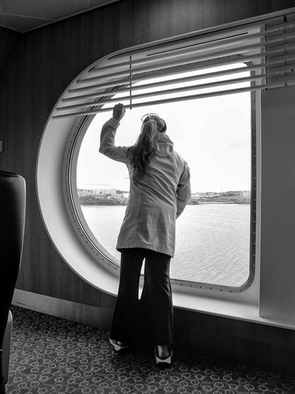 A person is standing in front of a large round window. Outside you can see the sea and a coastline.
***
Une personne se tient face à une grande fenêtre arrondie. Dehors on aperçoit la mer et une côte.