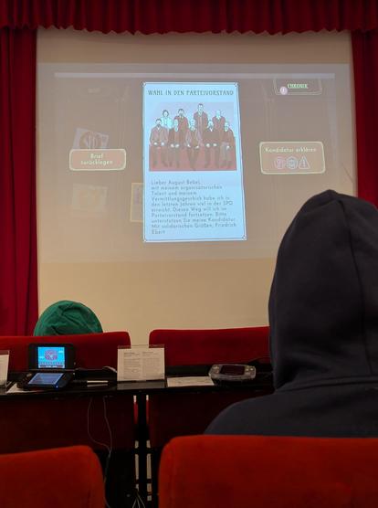 Menschen sitzen auf Kinositzen vor einer Leinwand. Die Leinwand ist in der Mitte des Bildes. Auf der Leinwand ist ein Videospiel. Verschiedene Elemente des Spiels sind am Rand, in der Mitte ist ein fiktiver Brief von Friedrich Ebert and August Bebel. Ebert bittet Bebel um Unterstützung bei Eberts Kandidatur zum Parteivorstand.