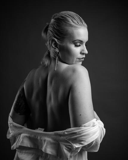 The image is a black-and-white photograph of a person with a bare back, partially covered by a white garment. They have short, styled hair, and they are wearing a long earring. The person is turned slightly to the side, looking downward. The lighting emphasizes the contours of their back and face, creating a dramatic effect.
