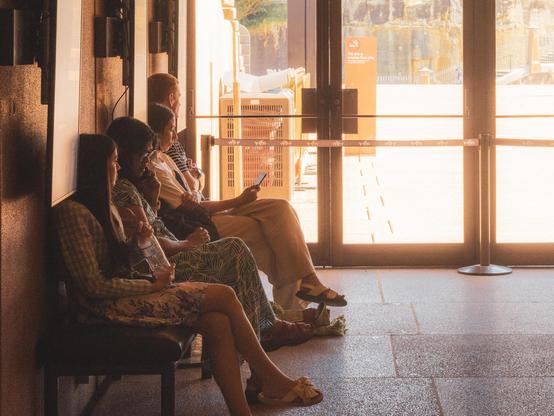 The image shows a group of people sitting on a bench indoors near a large glass door or window. They appear to be waiting, and one person is holding a phone. It’s a well-lit area with light coming in from outside.