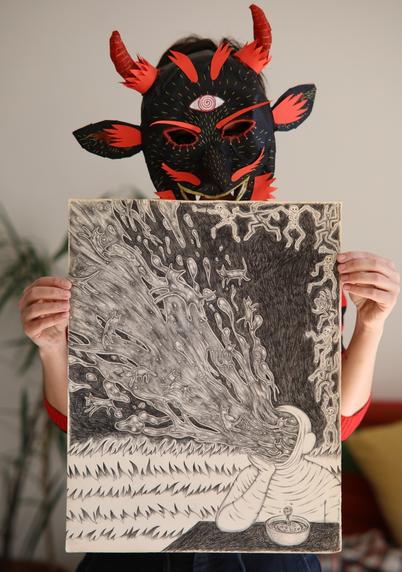 Photo d'une personne tenant un dessin de grand format. La personne porte un masque rouge et noir s'inspirant du personnage de Krampus, il est réalisé en papier.
Le dessin représente une personne appuyée sur une table dans le coin droit en bas. De son visage sort un flot d'un genre de liquide visqueux et étoilée où l'on distingue des silhouettes de chats. En dessous, l'horizon est composé de plusieurs étages de flammes. Sur les côtés formant le coin haut droit on peut voir des silhouettes de démons et de squelettes qui dansent, aligné•es. 
Le dessin est réalisé à la pierre noire, ça ressemble au crayon à papier en plus foncé.