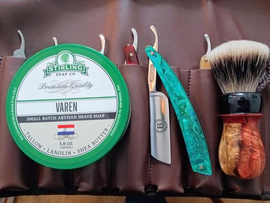 (Le Chat Mistral description)

The image showcases a traditional shaving set laid out on a leather surface. It features a green container of Stirling Soap Co.'s "Varen" shaving soap, a straight razor with a turquoise handle, and a badger hair shaving brush with a wooden handle. The soap container highlights ingredients like tallow, lanolin, and shea butter, emphasising quality and craftsmanship. The arrangement exudes a classic, refined aesthetic, ideal for a meticulous shaving routine.