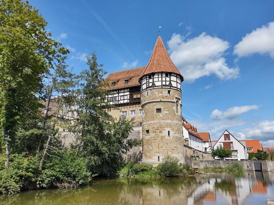 Das Bild zeigt ein historisches Schloss mit einem markanten runden Turm, der ein spitzes, rotes Ziegeldach trägt. Der Turm ist aus Naturstein gebaut und im oberen Bereich mit Fachwerk versehen, was ihm ein traditionelles und charmantes Aussehen verleiht. Das Hauptgebäude hinter dem Turm weist ebenfalls eine Mischung aus Steinmauerwerk und Fachwerk auf. Im Vordergrund ist ein ruhiger Fluss zu sehen, der die Burganlage spiegelt. Üppige grüne Vegetation, darunter Bäume und Sträucher, umrahmt das Gewässer und verleiht der Szene eine natürliche Atmosphäre. Der Himmel ist blau mit einigen weißen Wolken, die für ein freundliches, sonniges Wetter sprechen. Im Hintergrund sind weitere Fachwerkhäuser mit roten Dächern zu erkennen, die zur idyllischen, historischen Kulisse beitragen.