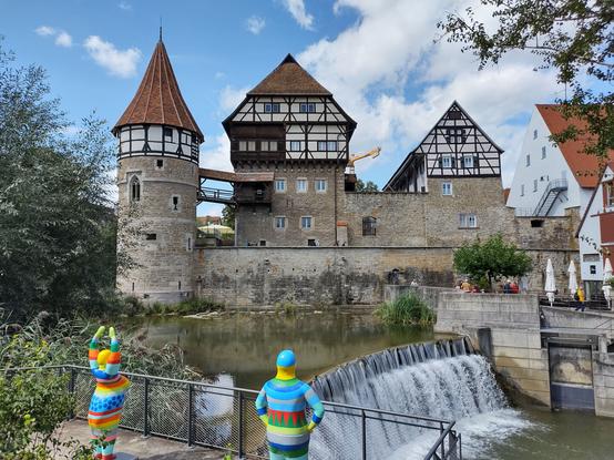 Das Bild zeigt eine malerische historische Szenerie mit einem Fachwerkgebäude, einem Wehrturm und einer Steinmauer, die an einem Fluss oder Kanal liegt. Eine kleine Staustufe erzeugt einen Wasserfall, der in ein ruhiges Gewässer mündet. Im Vordergrund stehen zwei bunte, moderne Skulpturen in abstrakter menschlicher Form. Die Fachwerkhäuser und der Turm verleihen der Szenerie einen mittelalterlichen Charme, während die Skulpturen einen modernen Kontrast darstellen. Der Himmel ist blau mit einigen weißen Wolken, und es sind einige Menschen zu sehen, die die Umgebung genießen.