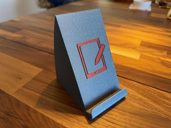 a photograph of the tablet stand on a wooden table. It's been printed in sparkly grey filament with a burgundy red icon of a tablet and stylus in the center.