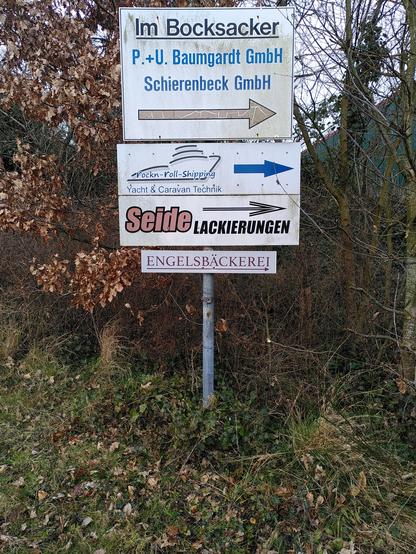 Vor verschiedenen Bäumen mit teils braunen Blättern vom Vorjahr steht ein Pfeiler mit drei Geschäftsschildern. Das unterste Schild weist auf mein Café hin: Engelsbäckerei (darunter ist ein Pfeil, der nach rechts weist).