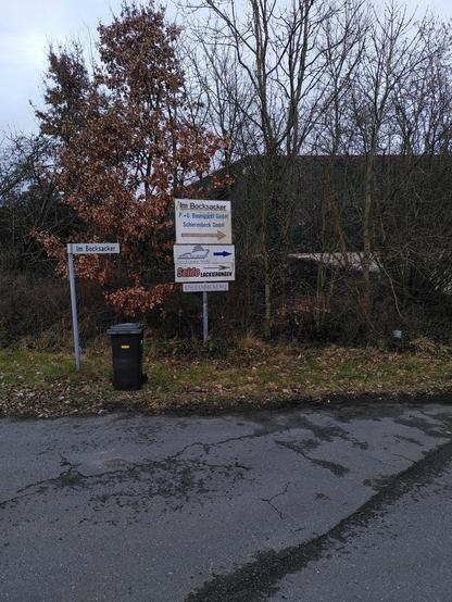 Ein Blick auf die Straßenecke *Im Bocksacker* im Gewerbegebiet von Bramstedt in Hagen im Bremischen. Vor verschiedenen Bäumen mit teils braunen Blättern vom Vorjahr stehen zwei Pfeiler aus Metall. Der linke trägt ein Straßenschild mit der Aufschrift *Im Bocksacker*, der daneben beherbergt drei Geschäftsschilder. Das unterste verweist auf mein Café Engelsbäckerei.