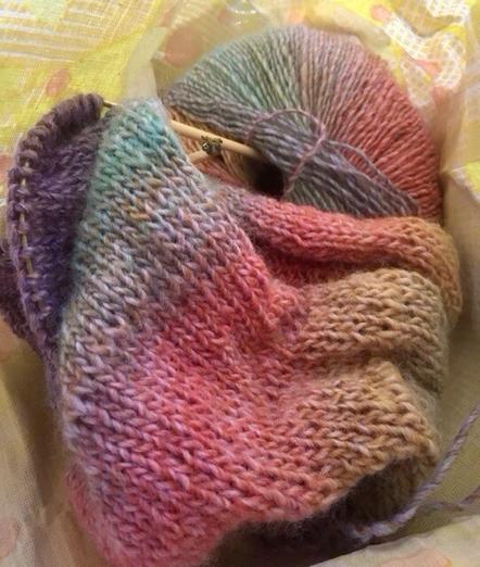 Foto bearbeitet. Ein Knäuel Farbverlaufsgarn, in dem zwei Stricknadel enden stecken, an einer hängt ein winziges Faultier. Weiterhin ist ein Stück des bunten Strickwerks zu sehen, „glatt rechts“ gestrickt.