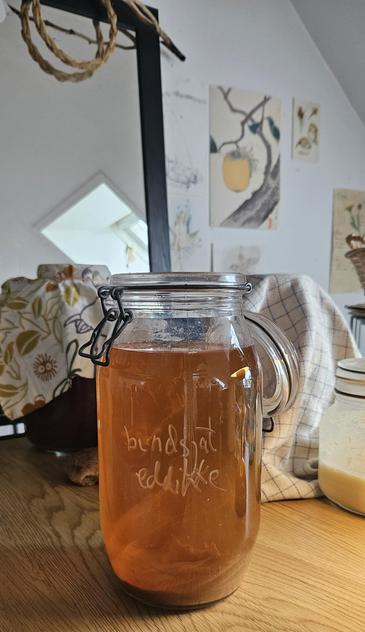 Jars covered in cloths in the background. Large open jar with golden liquid, labelled with chalk marker on the side "bundsjat eddikke"
Mirror reflecting a Sunny window in the background. Art on the wall.