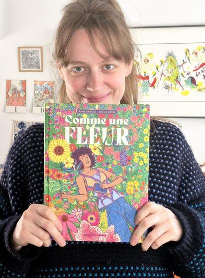 This is a photo of me facing the camera, holding the graphic novel Comme une fleur in a way that showcases its cover. On it, we see the main character, Églantine, lying amidst a field of flowers, a mix of lupins, sunflowers, poppies, and many more… The book will soon be available in bookstores, and I’ve just received the very first copy! JOY! This graphic novel, written by Anne Baraou and illustrated by me, will be released on March 19 by Delcourt.