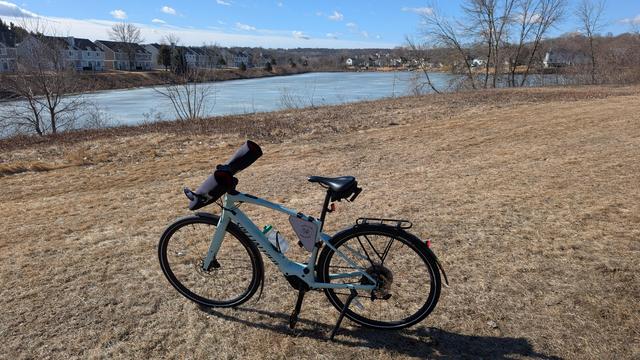 Sage green Vado SL ebike with Bar Mitts parked off the Cascade Lake Trail looking Southwest toward Interlachen Lake. There is brown lawn in the foreground, the lake is frozen over and there are townhomes on the far shore. The sky is mostly blue.