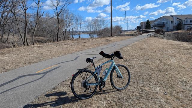 Sage green Vado SL ebike with Bar Mitts parked off the Cascade Lake Trail looking East back toward Manorwoods Lake. The asphalt trail heads back to a bridge over Cascade Creek which links the manmade lakes and there are townhomes behind on the right, utility poles center and bare trees left. The sky is blue with puffy white clouds.