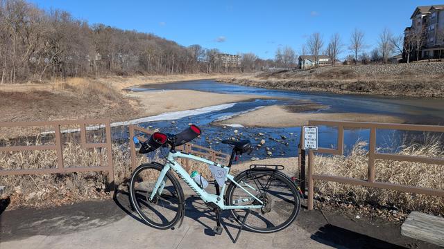 Sage green Vado SL ebike with Bar Mitts parked off the Cascade Lake Trail looking North at the confluence of Cascade Creek and the Zumbro River. There are bare trees on the left and senior living apartments on the far right. The sky is blue.