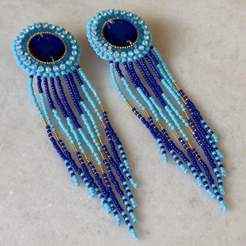 A pair of flat beadwork earrings with fringe on a marble background. The top of the earrings have royal blue rhinestone centers, which are outlined with tiny gold seed beads, blue satin beads, blue rhinestone banding, and larger blue seed beads on the edge. The fringe is royal blue to sky blue, to gold, royal blue, and sky blue again.