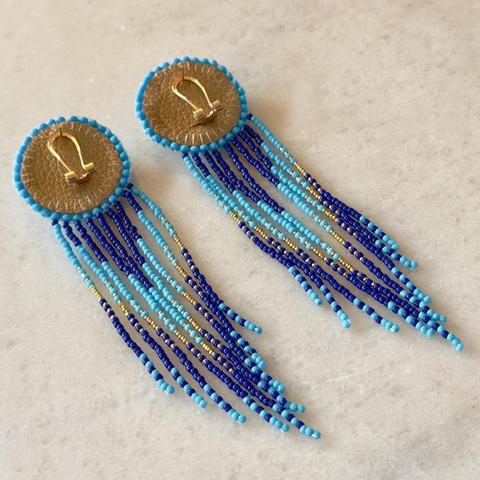 The same pair pair of flat beadwork earrings with fringe upside down on a marble background. The top of the earrings feature gold fingernail posts and a sand colored faux leather backing with blue seed beads stitched around the edge.