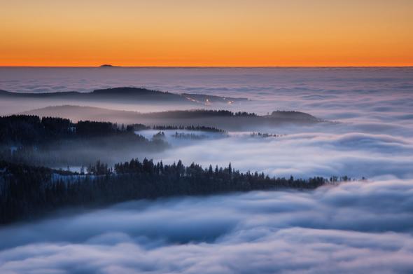 Blick über ein Nebelmeer bis zum Horizont in der Abenddämmerung. Einige bewaldete Höhenzüge ragen von links ins Bild und versinken nach rechts im Nebel. Das Bild besteht überwiegend aus blauen Farbtönen, während der Himmel kurz nach Sonnenuntergang orange leuchtet. Der Grand Ballon in den Vogesen ist klein am Horizont über dem Nebel zu erkennen.