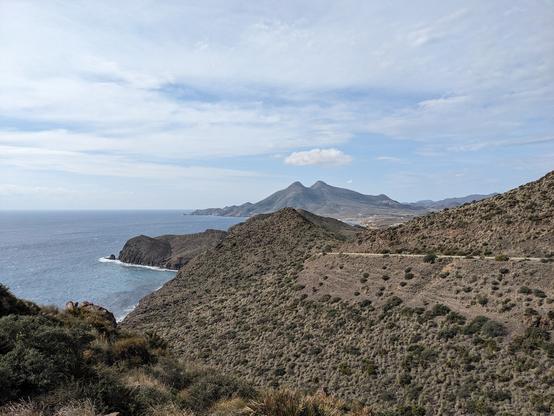 Brown, scrubby mountains before a rocky, wild coast with blue sea and white, crashing waves