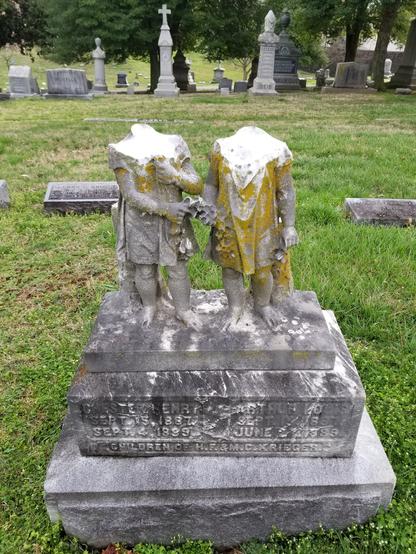 Gravestone, statuary of two standing children, heads missing. These twins died in 1899 at age two.