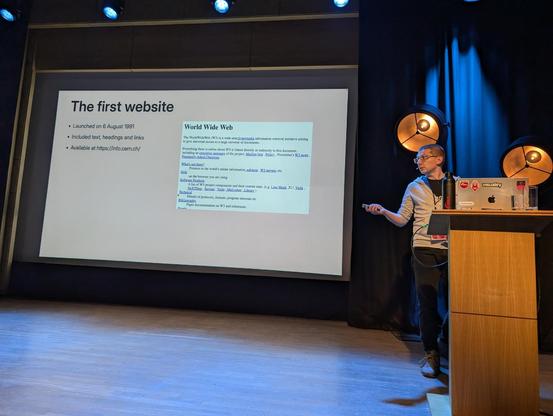Pawel on stage with a slide saying "The first website"