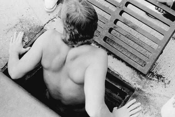 Black and white photo of a shirtless man lifting himself out of a street sewer. He is seen from the back, his hands on either side of the sewer opening, his shoulder blades pressed together, and the back of his shaggy mullet hairstyle. A hand from someone else can be seen holding up the sewer grate cover for him.