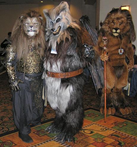 Three fursuiters in a hotel hallway - a lion in fancy black and gold patterned armor, mane braided under their chin; a brown and grey werewolf wearing a tooled leather belt; and a tan manticore with his wings folded behind him, holding a skull-topped staff and wearing nothing but his con badges.